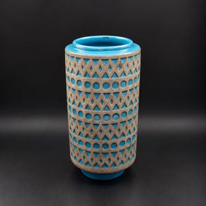 May include: A cylindrical ceramic vase featuring a vibrant turquoise blue glaze on the interior and rim, contrasted by a textured beige exterior surface adorned with a repeating geometric pattern of recessed circles and triangles.