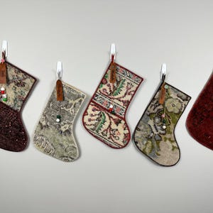 Puede incluir: Cinco calcetines navideños colgados en fila, cada uno confeccionado con tela estampada en varios tonos de rojo, verde, beige y marrón. Cada calcetín presenta una pequeña etiqueta de cuero marrón con un nombre y un dije de cuentas decorativo.