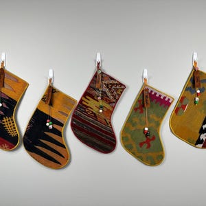 May include: Five decorative stockings hanging in a row, each crafted from woven fabric with distinct geometric patterns in shades of mustard yellow, deep red, black, and olive green. Each stocking features a leather tag and small colorful beads.