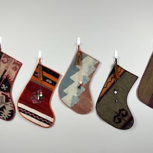 May include: Five hanging Christmas stockings made from woven kilim fabric, each featuring distinct geometric patterns in colors including red, orange, blue, green, and brown. Each stocking includes a leather tag and a small beaded charm.