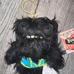 May include: A black plush keyring shaped like a fuzzy creature with green eyes, white teeth, and a light blue patterned waistband. A beige plastic ring is attached to the top, and a product tag with brick-patterned graphics is visible on the side.