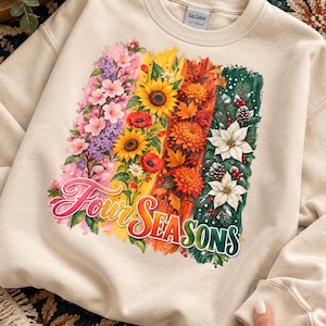 May include: A sand-colored crewneck sweatshirt featuring a graphic print of four vertical panels representing the seasons. The design includes pink cherry blossoms, yellow sunflowers, orange autumn leaves, and green pine branches with white poinsettias. The text "Four SEASONS" is printed in a stylized, colorful font across the bottom of the graphic.