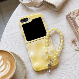 May include: A yellow, wavy-edged foldable phone case with a glittery finish. The case features a detachable wrist strap made of clear, round beads and a single, translucent yellow flower charm attached with a silver-toned metal lobster clasp.