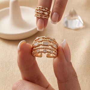 May include: A gold-toned multi-band ring featuring a combination of twisted metal textures, bezel-set round stones, and horizontal baguette-cut stones. The ring has an open-back design and is constructed from several interconnected horizontal bands.