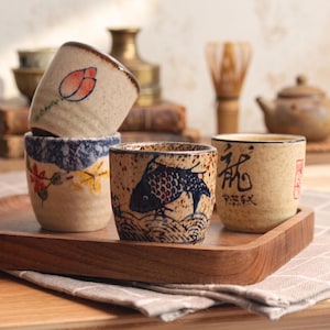 May include: Four ceramic tea cups with speckled beige surfaces. One cup features a painted orange flower, another displays blue and yellow autumn leaves, a third shows a dark blue fish, and the fourth cup has black Japanese calligraphy characters.