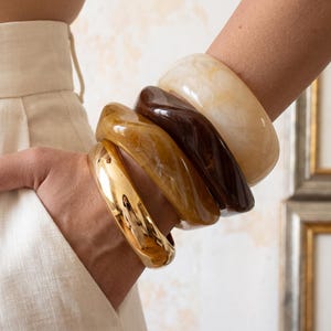 Puede incluir: Un conjunto de cuatro brazaletes apilados en la muñeca. La colección incluye un brazalete de metal dorado pulido, un brazalete de resina veteada en color crema translúcido, un brazalete de resina veteada marrón oscuro y uno de color ámbar.