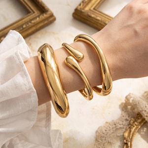 Puede incluir: Un conjunto de dos pulseras de metal dorado con un acabado liso, pulido y reflectante. Una pulsera es un brazalete sólido, grueso y curvado, mientras que la otra tiene un diseño más fino y abierto con extremos redondeados en forma de gota.