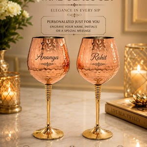 May include: A set of two hammered copper wine glasses with gold-toned stems and bases. Each glass features a personalised name, Ananya and Rohit, engraved on the bowl. The text reads Hammered Copper Wine Glass Set, Elegance in Every Sip.