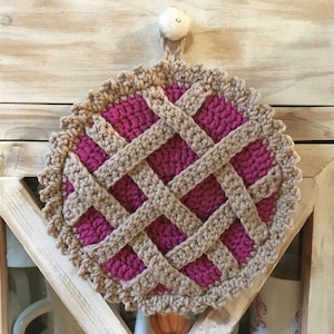 May include: A handmade crochet potholder shaped like a pie. The potholder features a magenta filling, a tan lattice crust, and a scalloped edge. It is hanging from a wooden cabinet with a white knob.