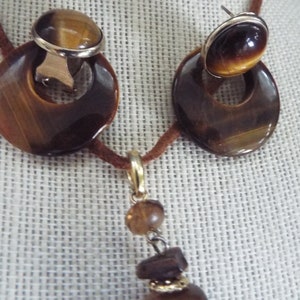 May include: A brown leather cord necklace with a tiger's eye pendant and matching earrings. The pendant is a round stone with a gold setting and the earrings are round stones with gold settings.