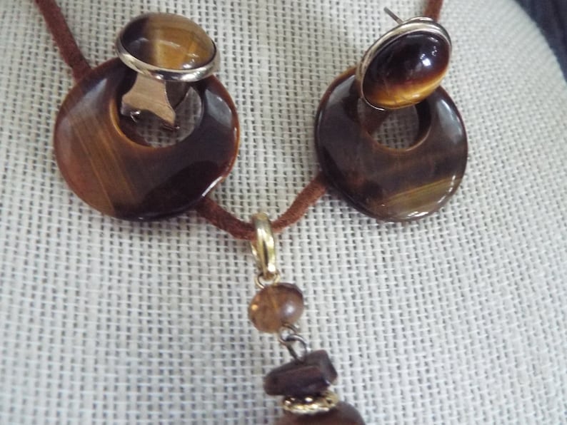 May include: A brown leather cord necklace with a tiger's eye pendant and matching earrings. The pendant is a round stone with a gold setting and the earrings are round stones with gold settings.