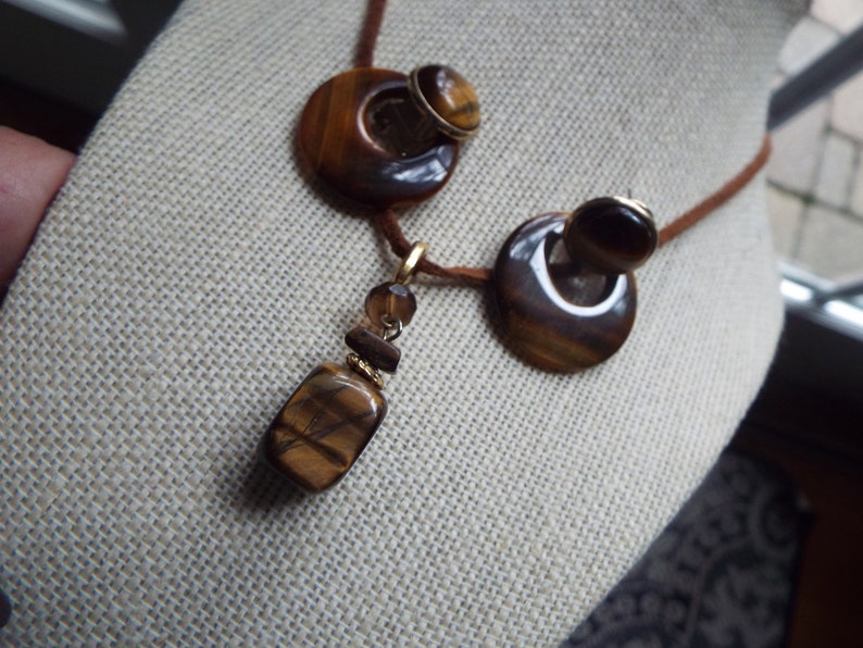 May include: A brown leather cord necklace with a large round tiger's eye pendant and matching earrings. The pendant is made of three stacked tiger's eye stones, the largest of which is rectangular.
