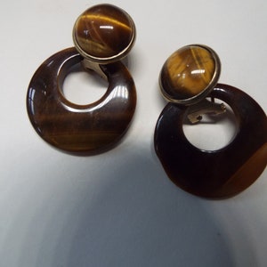 May include: A pair of gold tone clip-on earrings with tiger's eye stone accents. The earrings feature a round stone set in a gold tone bezel and a large, round, tiger's eye stone disc with a hole in the center.