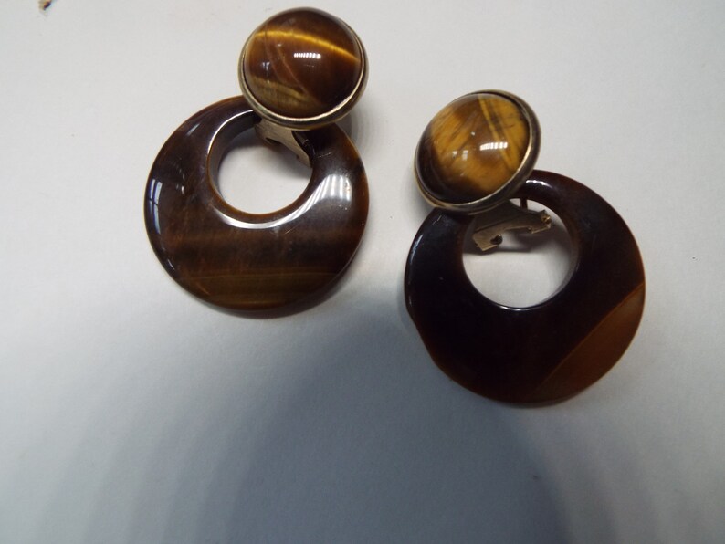 May include: A pair of gold tone clip-on earrings with tiger's eye stone accents. The earrings feature a round stone set in a gold tone bezel and a large, round, tiger's eye stone disc with a hole in the center.
