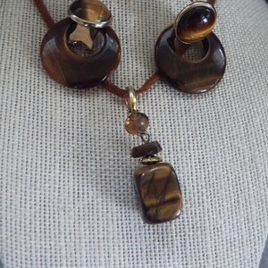 May include: A brown tiger's eye stone necklace and earrings set. The necklace features a long pendant with a rectangular tiger's eye stone, a smaller round brown stone, and gold accents. The earrings are clip-on style with round tiger's eye stones.