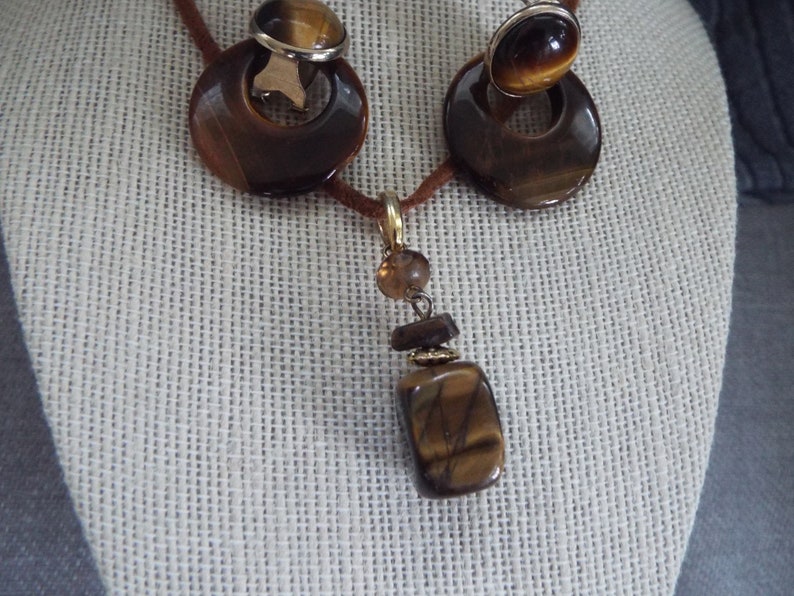 May include: A brown tiger's eye stone necklace and earrings set. The necklace features a long pendant with a rectangular tiger's eye stone, a smaller round brown stone, and gold accents. The earrings are clip-on style with round tiger's eye stones.