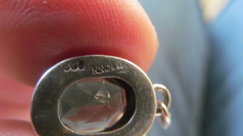 May include: Close-up of a silver pendant with a clear gemstone. The pendant has the inscription "925" and "NORWAY" on the back.