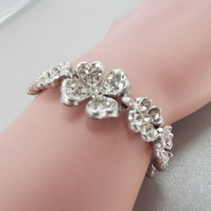 May include: A silver bracelet with a floral design. The bracelet is made up of several small flowers, each with a center stone and surrounded by smaller stones. The bracelet is on a mannequin's wrist.