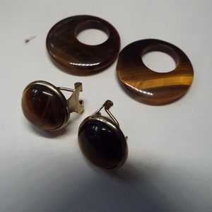 May include: A set of three tiger's eye earrings. Two of the earrings are round and have a hole in the center. The third earring is oval shaped and has a gold clasp.