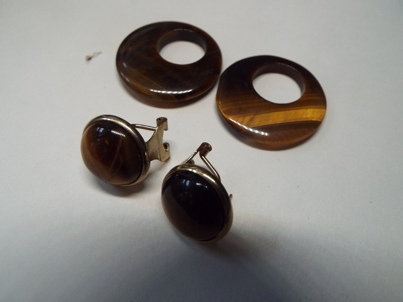 May include: A set of three tiger's eye earrings. Two of the earrings are round and have a hole in the center. The third earring is oval shaped and has a gold clasp.
