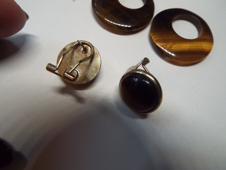 May include: A pair of clip-on earrings with tiger's eye stones. One earring has a round, dark brown stone, while the other has a round, gold-toned setting with a tiger's eye stone.