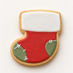 May include: A stocking-shaped sugar cookie decorated with smooth red icing in the center, a white icing cuff at the top, and two sage green icing patches on the heel and toe, each detailed with small white icing stitches.