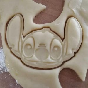 Pode incluir: Um cortador de biscoitos de plástico pressionado numa massa crua de cor clara, criando uma impressão detalhada da personagem Stitch. O cortador apresenta orelhas grandes, olhos largos e um nariz distinto, deixando um contorno claro na massa.