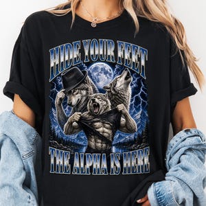 May include: A black t-shirt featuring a graphic print of three wolves, one wearing a fedora, with a full moon and lightning in the background. The text "HIDE YOUR FEET" is printed above the wolves, and "THE ALPHA IS HERE" is printed below.