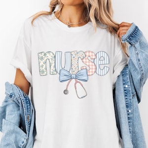 May include: A white short-sleeved t-shirt featuring the word nurse in patterned letters with a light blue bow and a stethoscope graphic underneath. The shirt is worn with a light blue denim jacket draped over the shoulders.