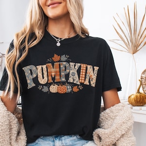 May include: A black short-sleeve t-shirt featuring the word PUMPKIN in large, multi-patterned block letters. The design includes various plaid and floral prints in orange, blue, and tan tones, accented by small illustrated pumpkins and leaves.