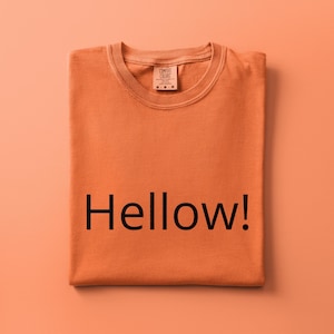 May include: A folded orange short-sleeve t-shirt with a crew neckline. The shirt features the black text Hellow! printed across the center. A Comfort Colors brand label is visible at the inner neckline, indicating a size medium for the garment.