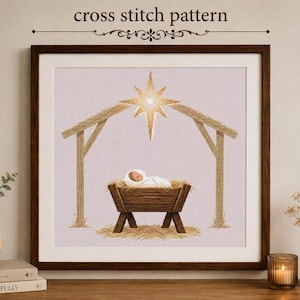 May include: A cross stitch pattern featuring a nativity scene with a baby in a wooden manger under a large golden star. The design is framed in a dark wood frame. The text "cross stitch pattern" is printed in black above the framed artwork.