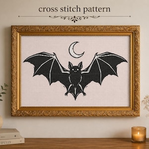 Può includere: Uno schema a punto croce raffigurante un pipistrello nero con le ali spiegate sotto una luna crescente. Il disegno è inserito in una cornice rettangolare dorata con bordo decorato, su una parete chiara con testo sovrastante.