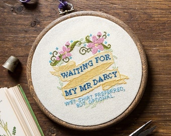 Op de afbeelding: Een borduurring met de tekst "Waiting for my Mr Darcy", versierd met bloemmotieven. De afbeelding toont ook een vingerhoed, een schaar, knopen en een vintage boek, allemaal op een houten oppervlak.