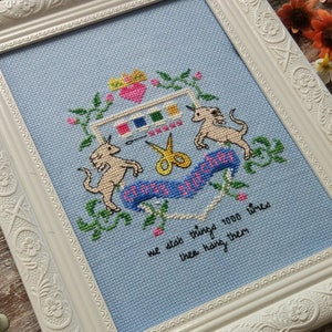 Może przedstawiać: Haftowany krzyżykiem obraz w białej ramie z niebieskim tłem przedstawiający dwa jednorożce, parę nożyczek i serce z tęczą i paletą farb. Tekst brzmi "Craft is Magic" i "We did a thing 1000 times then hang them".