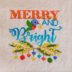 May include: Cross-stitch embroidery on a white canvas with the text "Merry and Bright" in colorful lettering. A small bird with a star is stitched above the text. A garland of yellow flowers with red, orange, blue, and green ornaments hangs below the text.