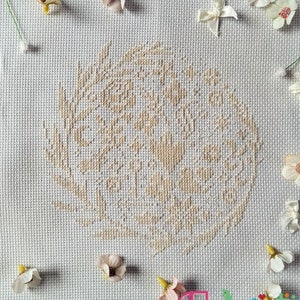 May include: A white cross-stitch embroidery with a beige floral design in a wreath shape. The embroidery is on a white fabric background.