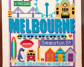 May include: A colorful cross-stitch design depicting various iconic landmarks and symbols of Melbourne, Australia. The design includes a Ferris wheel, a train, a surfboard, a penguin, a tram, beach huts, and the text "MELBOURNE" and "FOUR SEASONS IN ONE DAY".