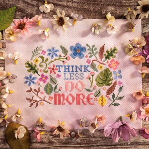 Puede incluir: Un bordado de punto de cruz rosa con un borde floral y el texto "Think Less Do More".