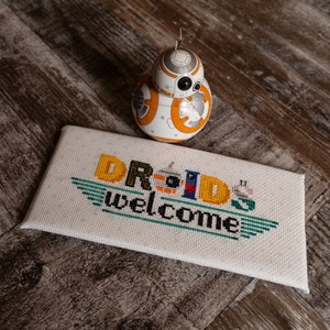 May include: A white and orange Star Wars BB-8 droid toy sits on a white canvas with the words "Droids welcome" embroidered in colorful thread.