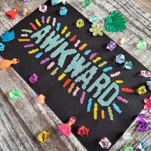 May include: A black canvas with the word "Awkward" stitched in colorful thread. The word is surrounded by colorful rays of light. The canvas is surrounded by colorful decorative items.