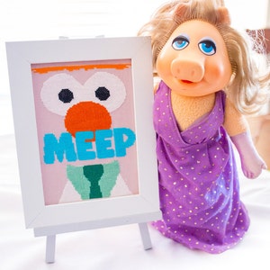 Könnte beinhalten: Ein weiß gerahmtes Kreuzstichbild mit rosa Hintergrund, das ein Comic-Gesicht mit großen Augen, einer orangefarbenen Nase und dem Wort "MEEP" in blauen Buchstaben zeigt. Eine Miss Piggy-Puppe in einem violetten Kleid mit goldenen Punkten steht neben dem gerahmten Kreuzstichbild.
