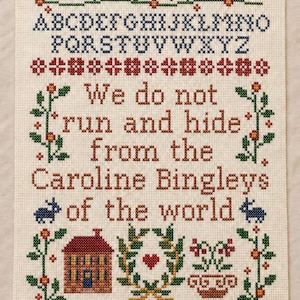 May include: A cross-stitch sampler on cream fabric featuring the alphabet in blue, a floral border, and the text: We do not run and hide from the Caroline Bingleys of the world. Includes motifs of a house, a heart in a wreath, and small animals.