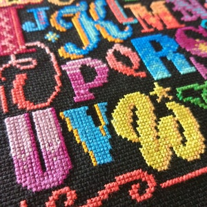 May include: A cross-stitch embroidery piece on black fabric featuring the English alphabet in various vibrant colors including pink, blue, orange, yellow, and green. Each letter is stitched with a textured thread pattern, creating a colorful display.