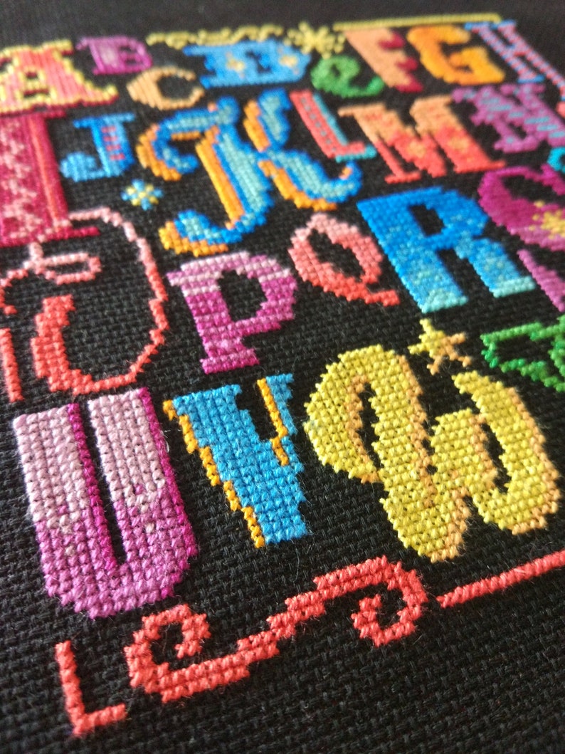 May include: A cross-stitch embroidery piece on black fabric featuring the English alphabet in various vibrant colors including pink, blue, orange, yellow, and green. Each letter is stitched with a textured thread pattern, creating a colorful display.