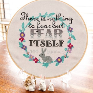 May include: A white embroidery hoop with the phrase "There is nothing to fear but FEAR ITSELF" stitched in black thread. The hoop is surrounded by a floral wreath with teal leaves and pink, purple, and yellow flowers. Three white ceramic bunnies are in the foreground.