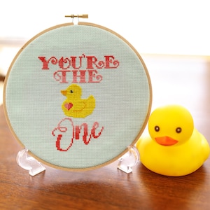 May include: A light blue embroidery hoop with the text "You're the one" in pink and red lettering. A yellow rubber duck with a red heart is in the center of the hoop.