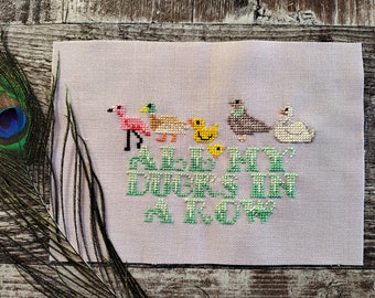 Puede incluir: Bordado de punto de cruz en tela rosa con un flamenco, patos, un pollito, una paloma y un cisne en fila. El texto dice "All my ducks in a row".