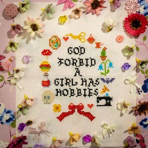 Op de afbeelding: Een kruissteekborduurwerk met de tekst "GOD FORBID A GIRL HAS HOBBIES", omringd door kleurrijke geborduurde afbeeldingen en decoratieve bloemen. De borduursel staat op een witte stof, omlijst door een bloemenpatroon.