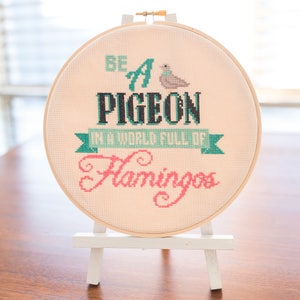 Könnte beinhalten: Ein beiger Stickrahmen mit weißem Hintergrund und schwarzem Text, der "Be a Pigeon in a World Full of Flamingos" lautet. Der Text ist in teal und rosa Schrift gehalten. Ein kleiner gestickter Vogel befindet sich über dem Text.